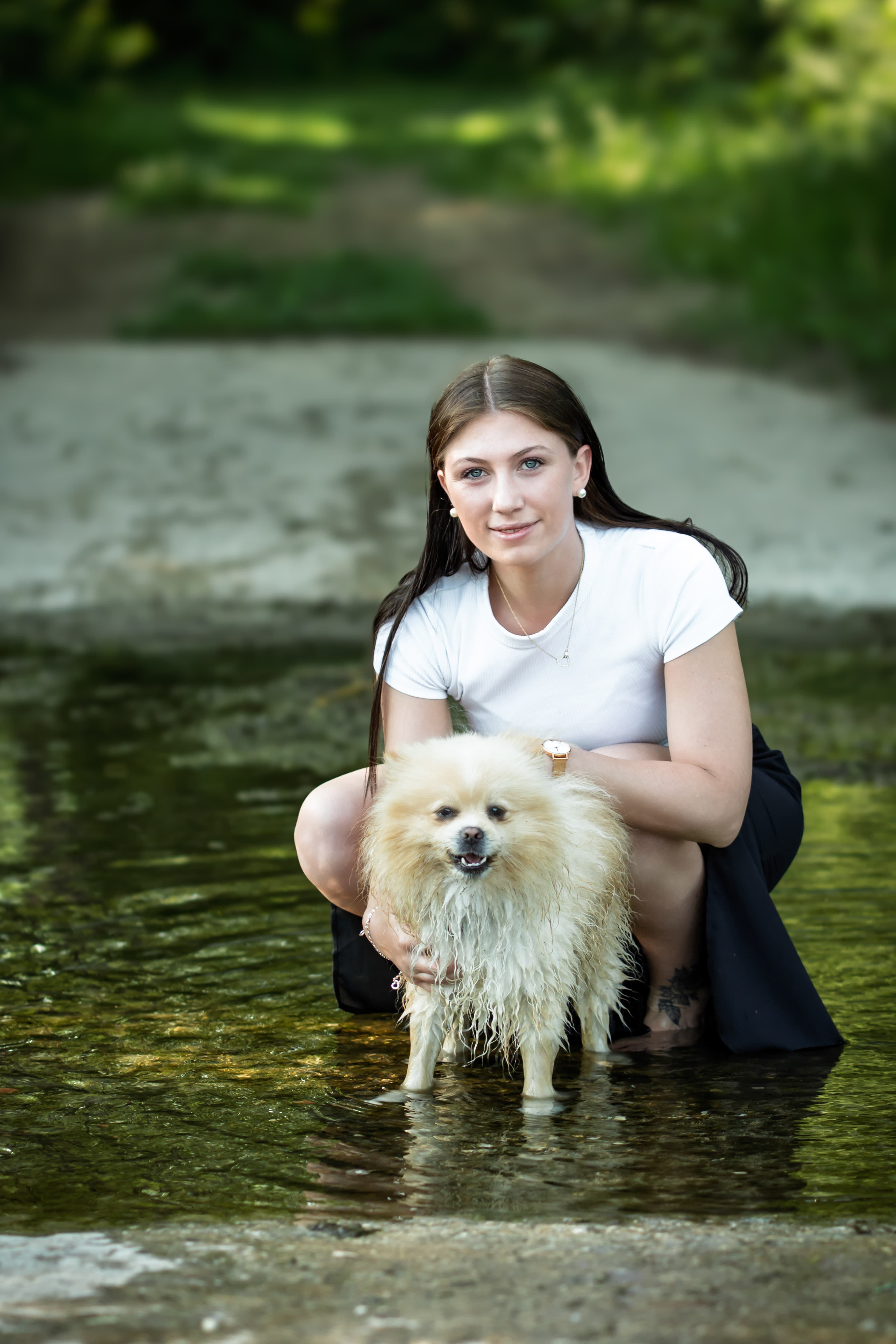 Frau kniet mit kleinem Hund im flachen Wasser, grüner Hintergrund mit Bokeh